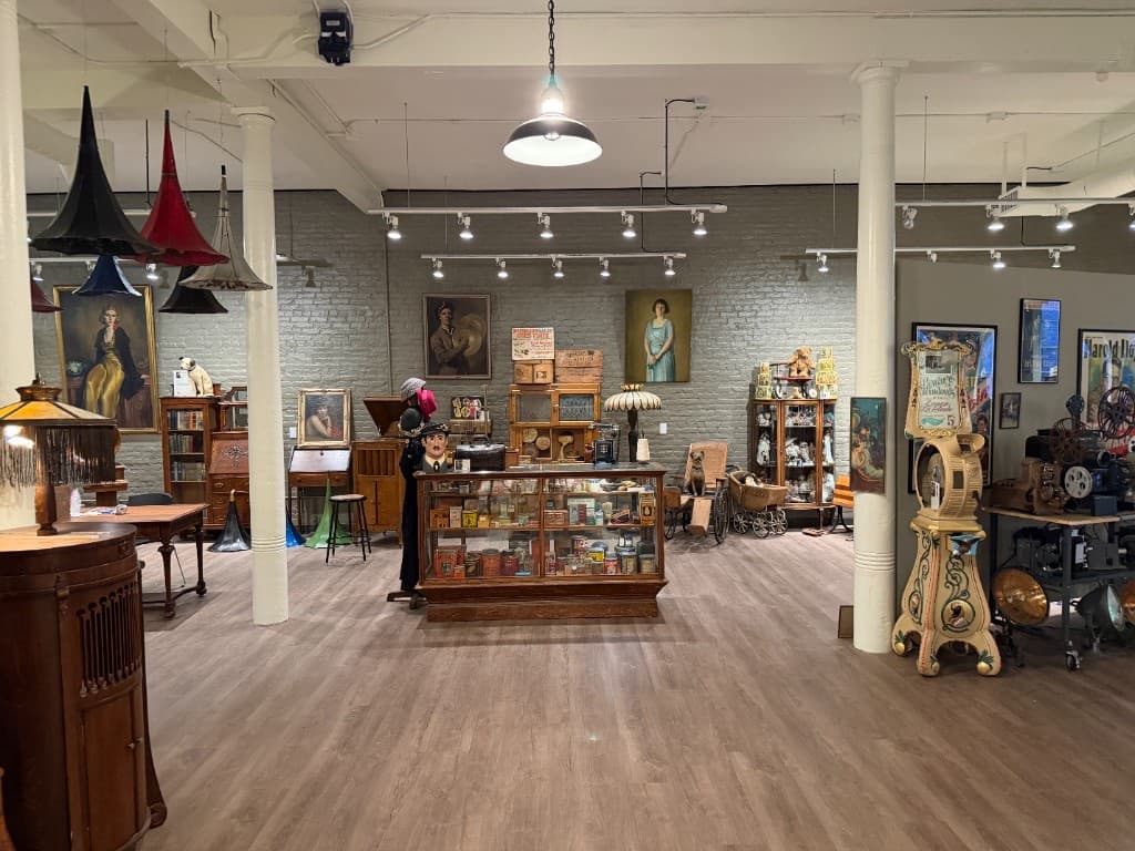 Wide gallery view with early film projectors and reels at right, paintings on the brick wall, and display cases across the room.