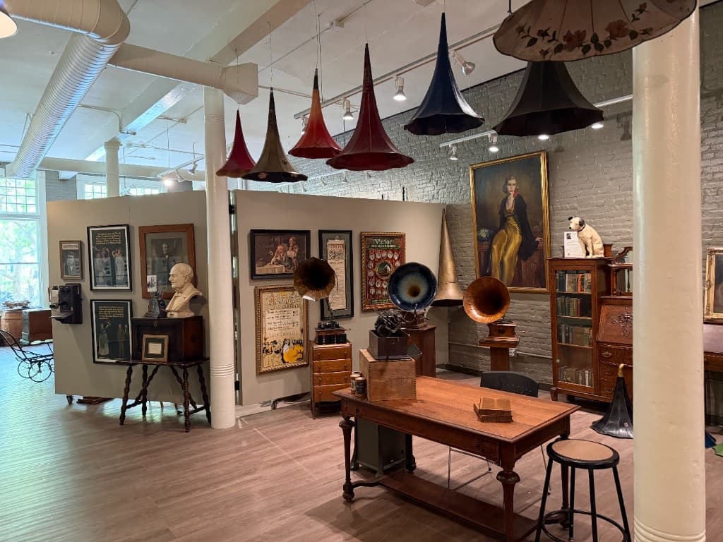 Several antique gramophones beneath a row of large suspended phonograph horns, with framed art and a portrait nearby.