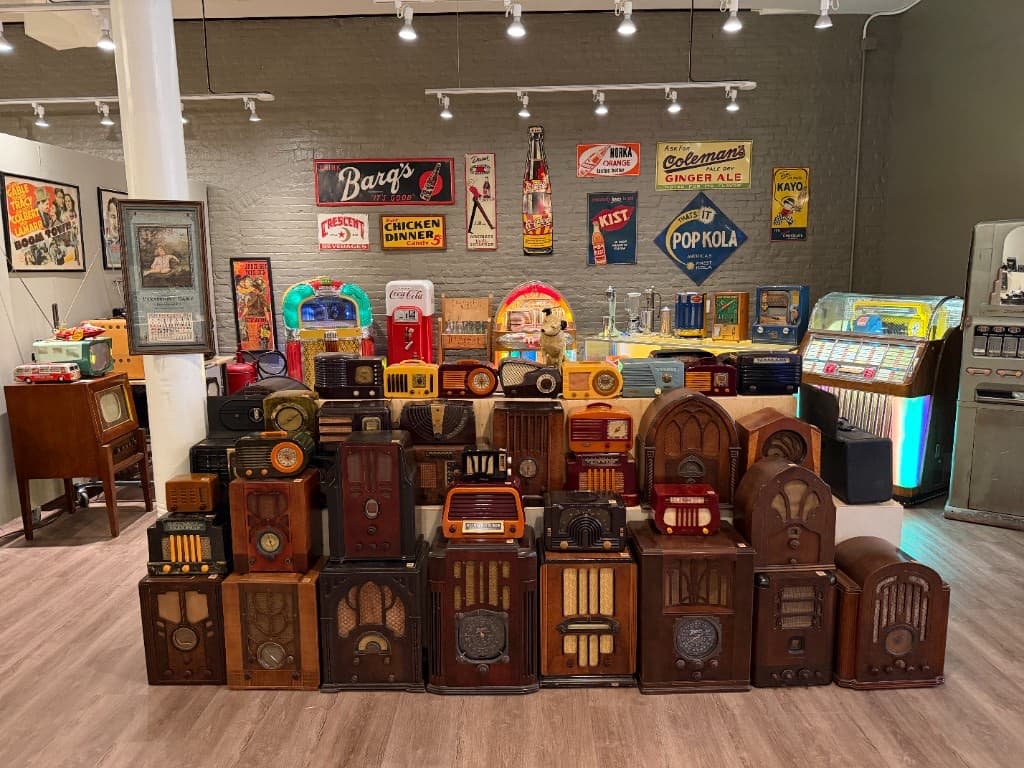 A dense display of dozens of vintage radios arranged below colorful soda signs, with jukeboxes behind them.